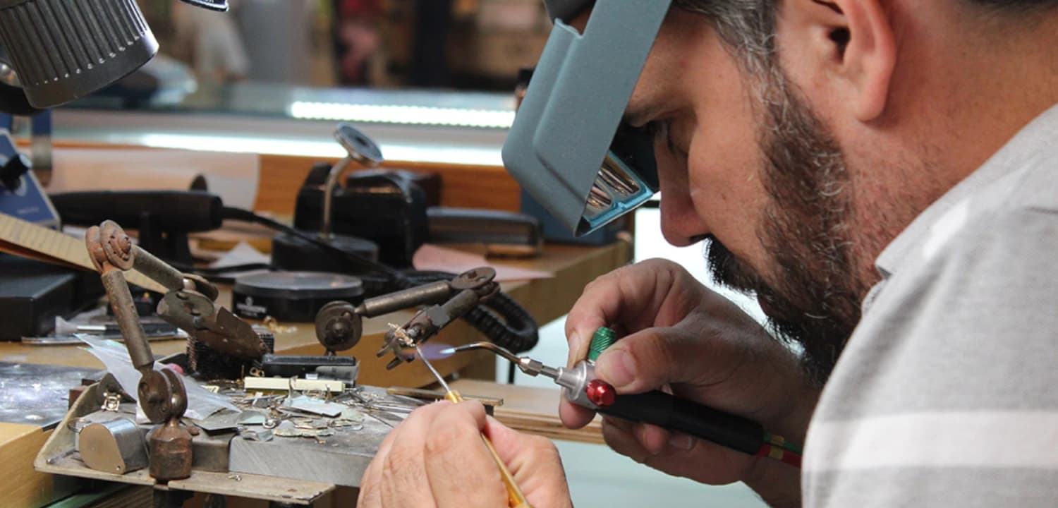Jeweler in white shirt soldering watch components at workbench with tools